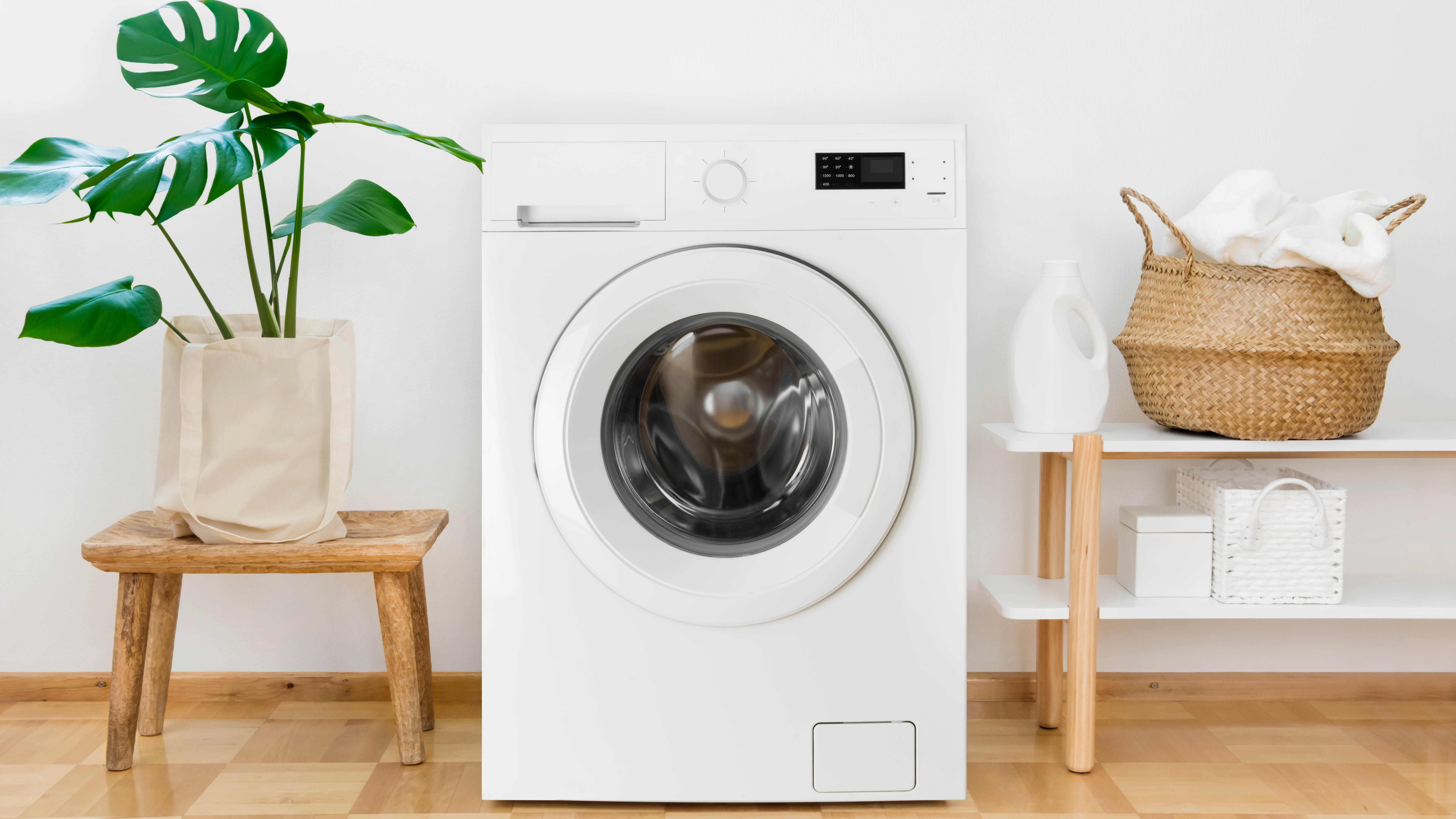 Une machine à laver à côté de la lessive, un panier à linge et une plante