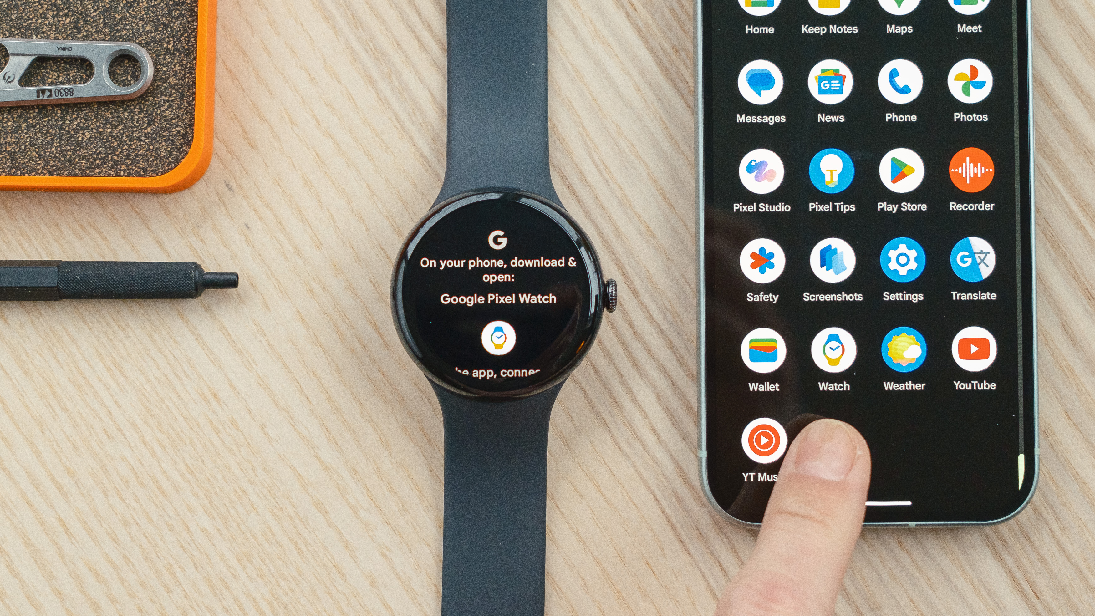 Google Pixel Watch 3 posée sur un bureau en bois à côté d'un téléphone Google Pixel 9, un doigt pointant vers l'application Watch