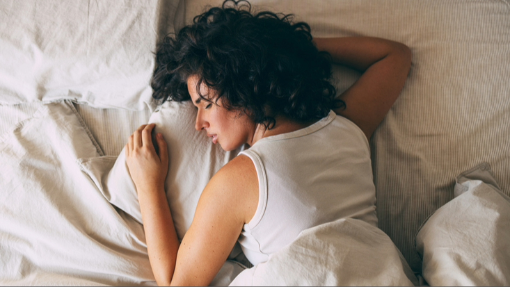 Femme aux cheveux noirs bouclés portant un débardeur blanc dormant sur le devant dans un lit avec des draps blancs.