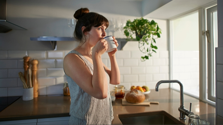 Une femme debout à la fenêtre avec une tasse de thé dans la cuisine avec une lumière du matin qui arrive.
