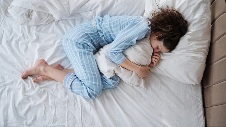Une femme portant un pyjama bleu dormant en position fœtale serrant un oreiller sur un lit avec des draps blancs.