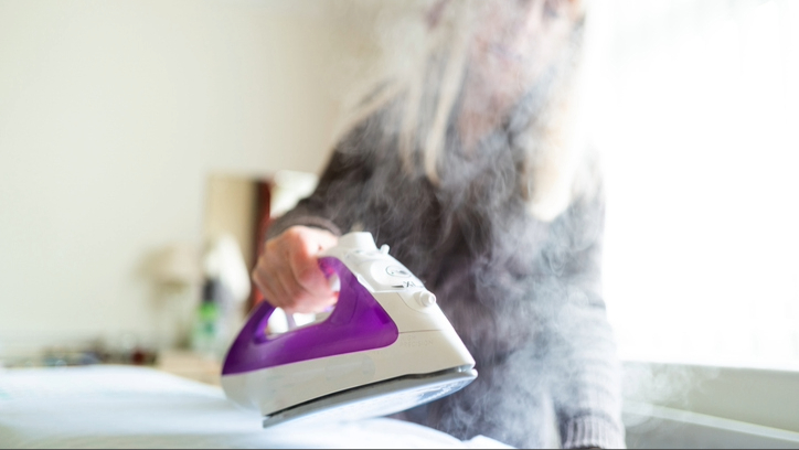 Une femme blonde tenant un fer à vapeur rose et blanc sur un lit blanc