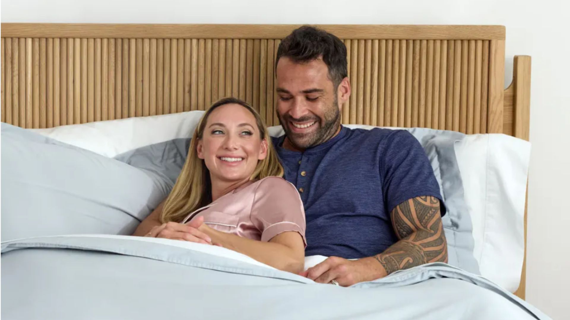 Un couple (la femme est blonde portant un pyjama rose et l'homme a les cheveux foncés et les tatouages et porte un haut bleu) au lit avec des draps bleus et une tête de lit en bois de rotin