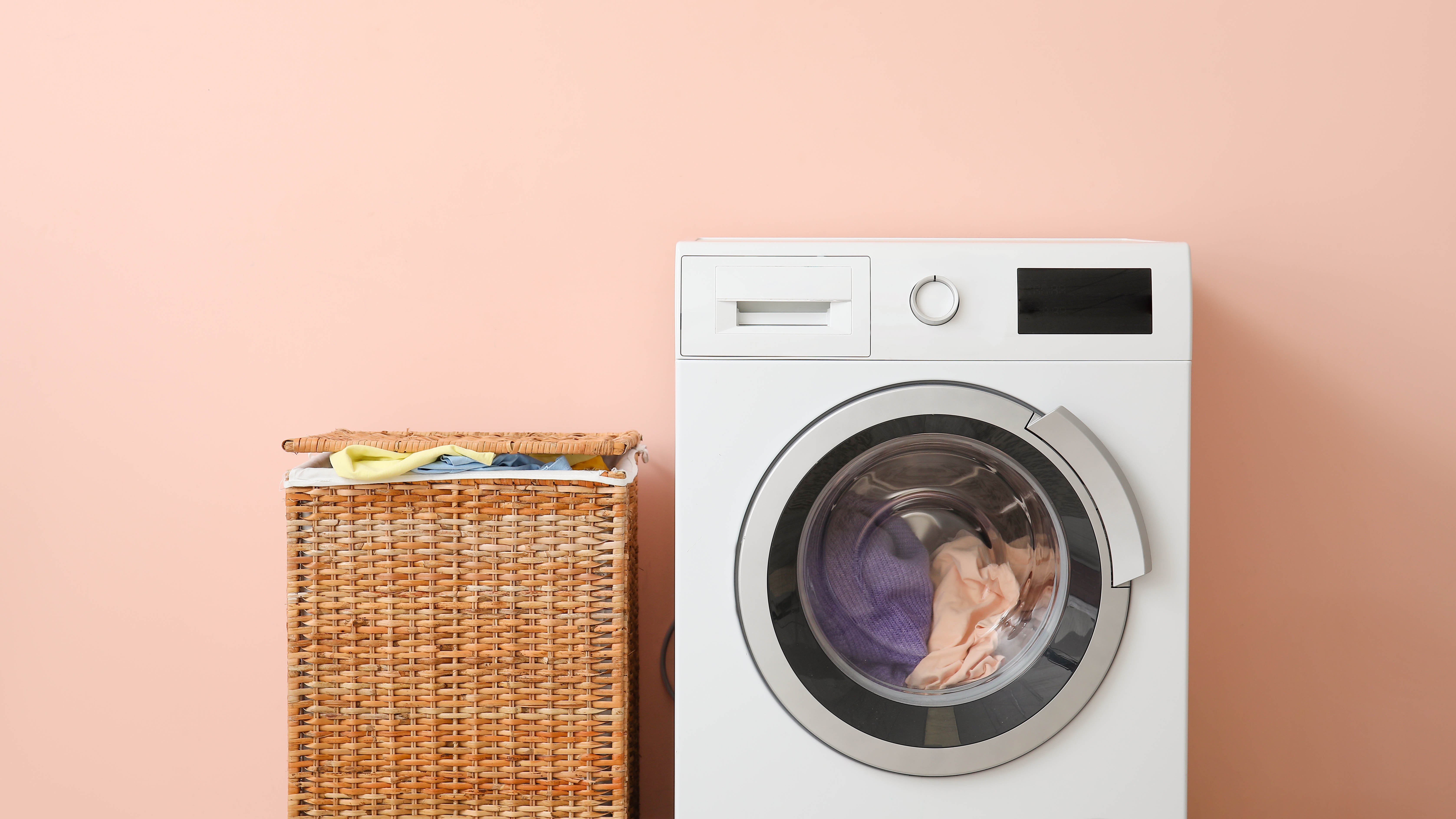 Une machine à laver avec des vêtements à l'intérieur à côté d'un panier à linge complet sur un fond rose