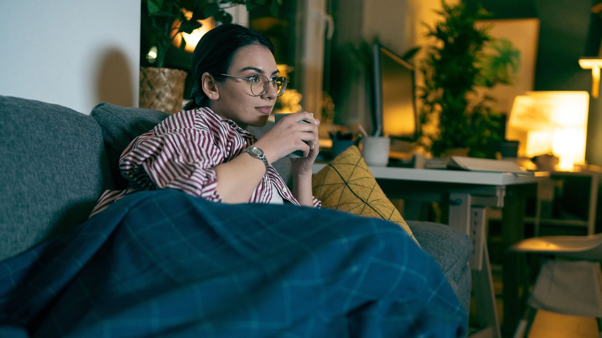 Une jeune femme aux cheveux noirs, portant des lunettes et sirotant une tasse de thé, regarde la télévision sur un canapé gris dans un salon faiblement éclairé.