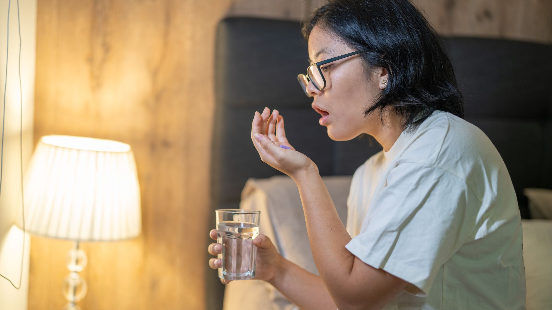 Une femme aux cheveux noirs portant des lunettes et tenant un verre d'eau est assise au bord de son lit et se prépare à prendre un supplément multivitaminé le soir.
