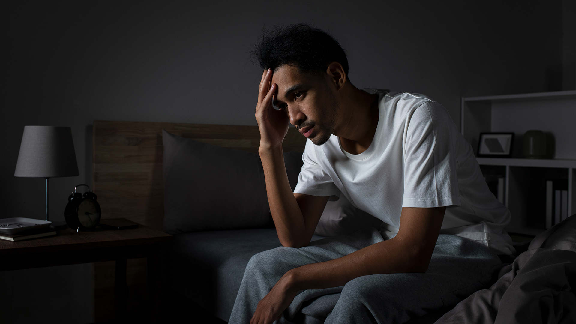 Un homme est assis sur le côté de son lit dans une pièce sombre, la tête dans les mains, car il éprouve de la détresse à cause de son incapacité à dormir.