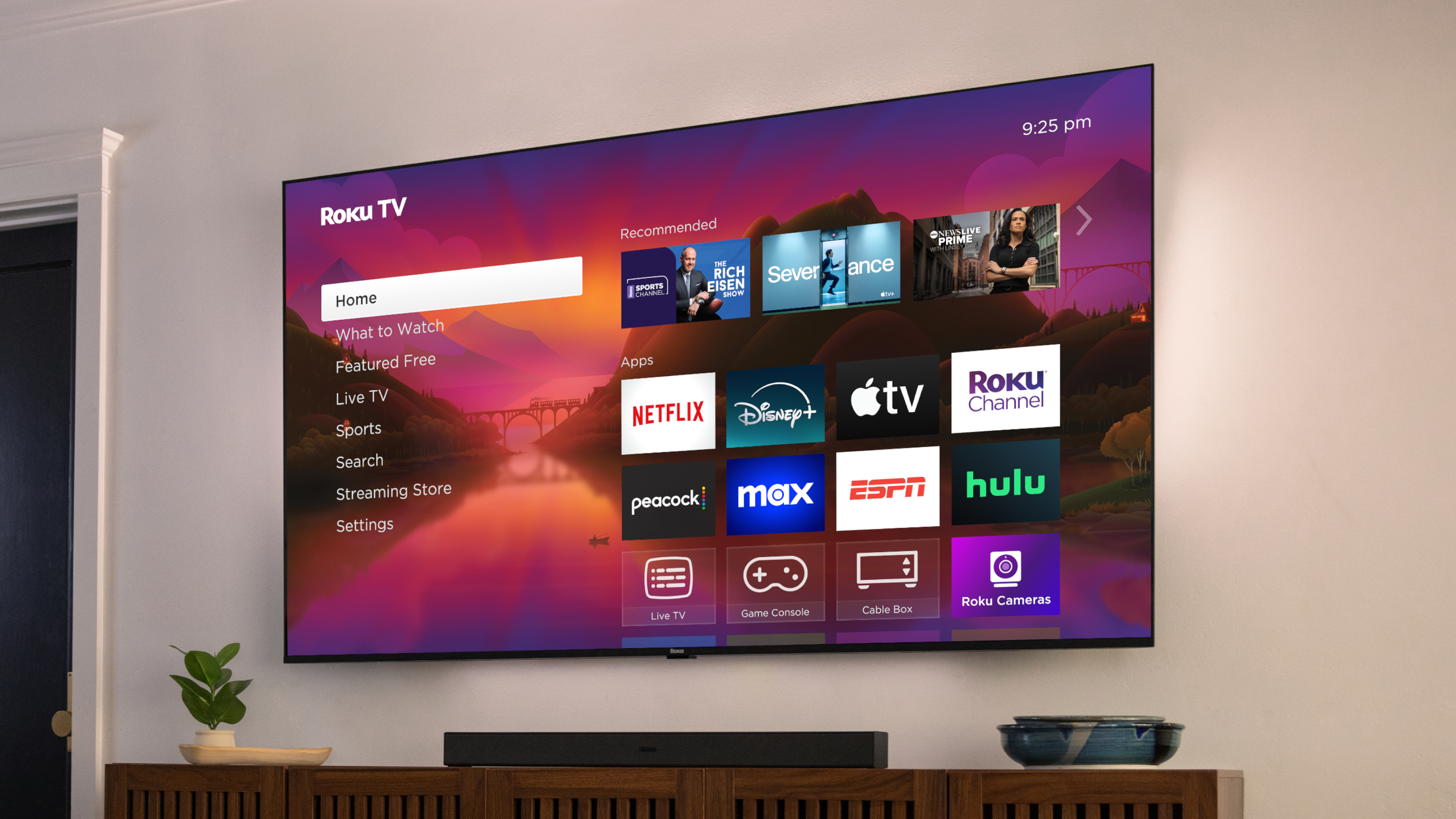 Le téléviseur Roku Plus Series 2025, fixé au mur dans un salon contemporain, affichant l'écran d'accueil Roku. L'écran d'accueil de Roku présente diverses applications de streaming.