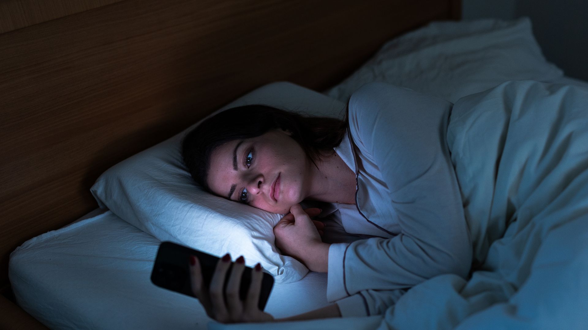 Une femme est allongée au lit la nuit et regarde son téléphone, elle est éclairée par la lumière de l'écran.