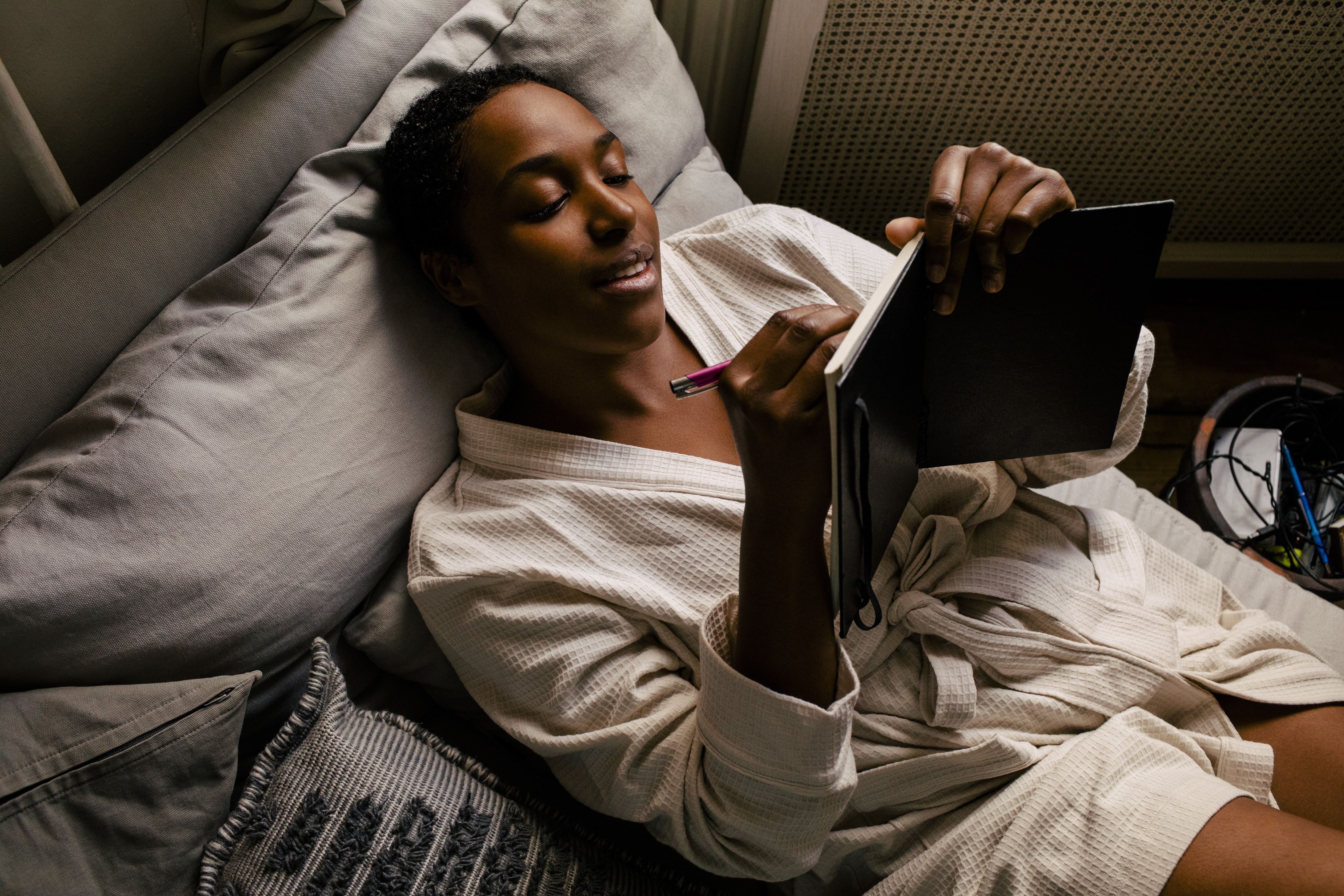 Une femme est allongée dans son lit, vêtue d'une robe de chambre, alors qu'elle écrit dans un journal.