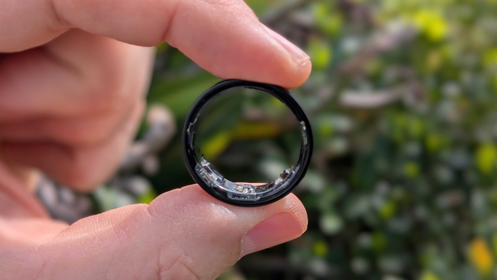 Photo en gros plan de la version noire du Galaxy Ring de Samsung, tenue entre le pouce et l'index devant un jardin.