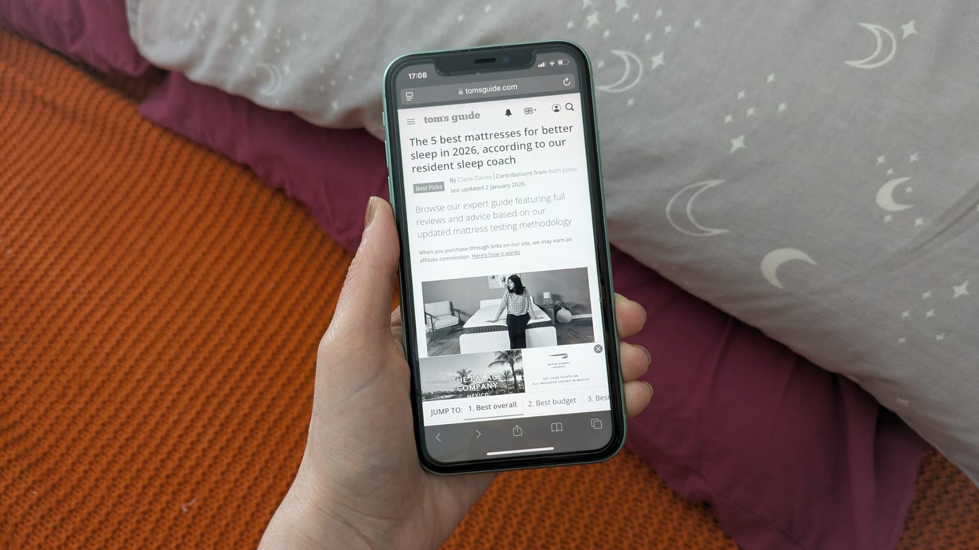 Une main tient un iPhone 11 montrant le guide du meilleur matelas Tom's Guide en niveaux de gris contre un lit