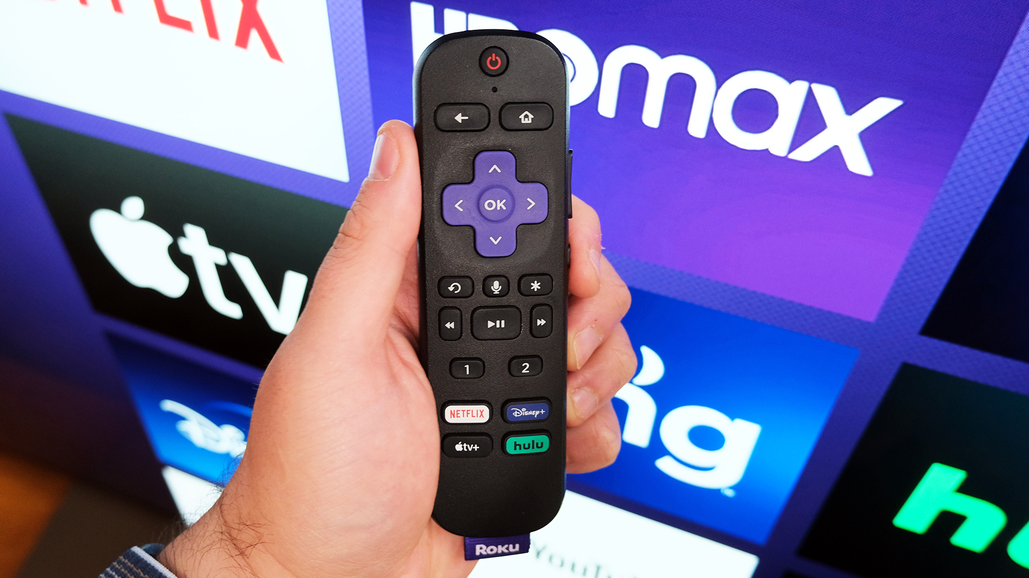 Une télécommande Roku à la main devant un téléviseur avec la page d'accueil de Roku.