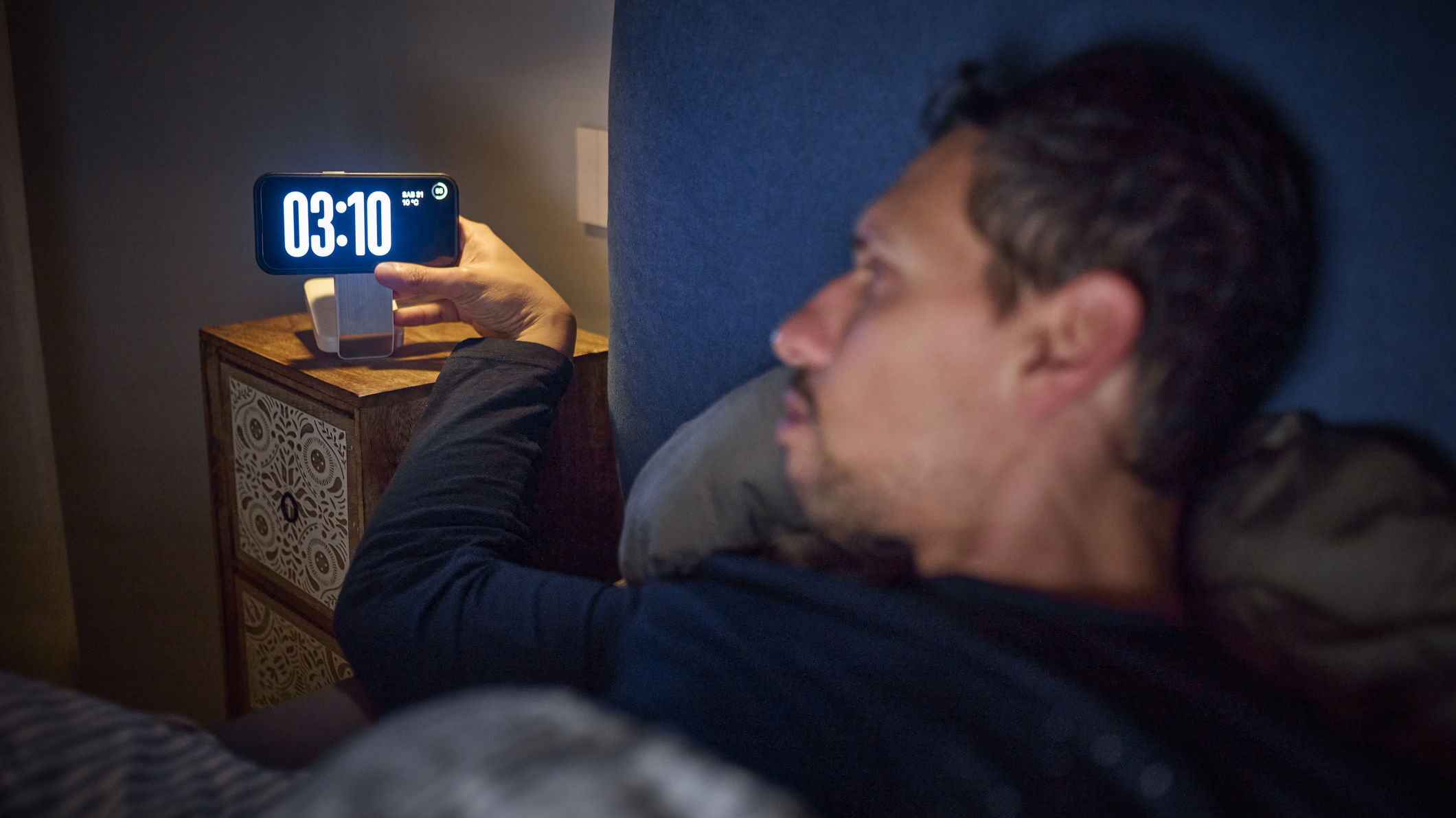 Un homme aux cheveux bruns courts se réveille au milieu de la nuit et commet l'erreur d'hygiène du sommeil en regardant son réveil, qui indique l'heure à 3h10.