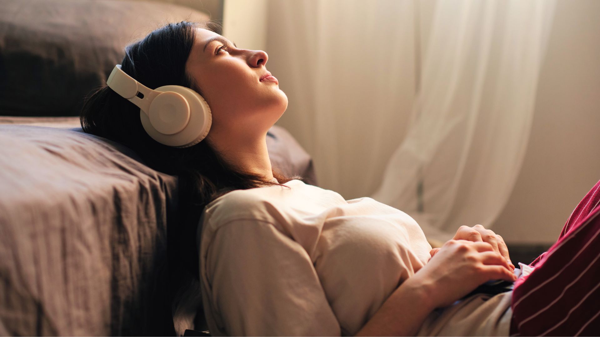 Une jeune femme brune se détend contre son lit tout en écoutant un podcast au casque.