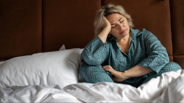 Une femme portant un pyjama bleu assise avec les jambes croisées et la tête appuyée sur la main dans son lit avec une literie blanche contre une tête de lit marron