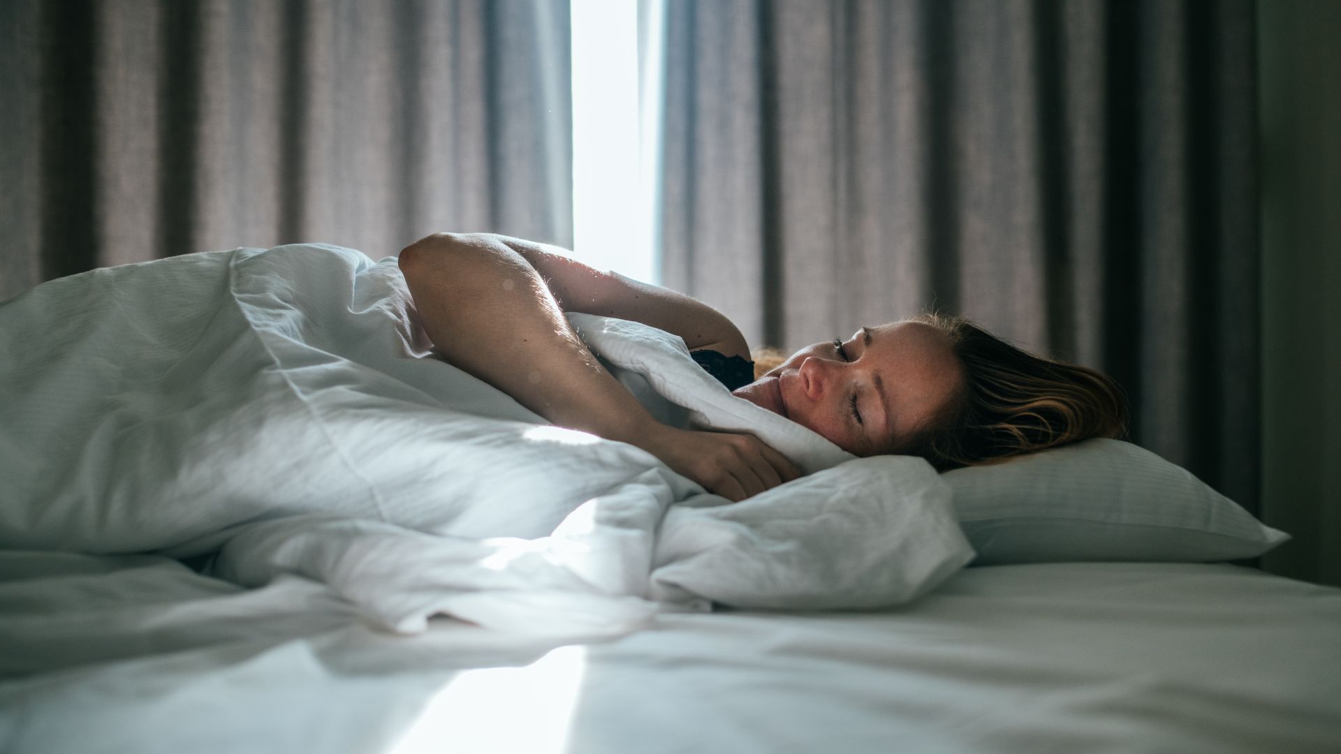 Une femme dort dans un lit recouvert de linge de lit blanc. En arrière-plan, des rideaux légèrement ouverts laissent entrer la lumière du soleil.